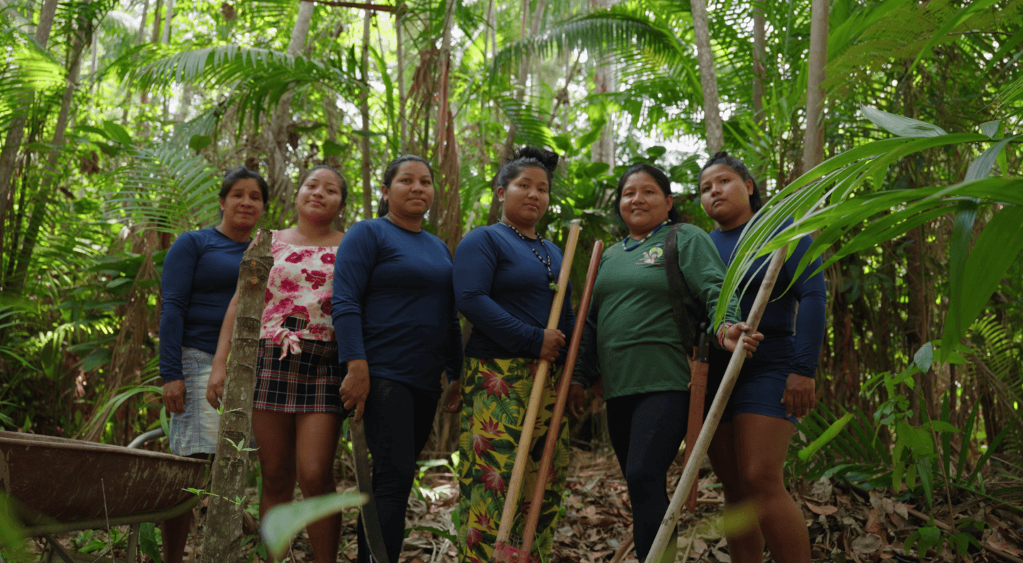 a group of Indigenous women stand together in the Amazon rainforest holding tools used for land restoration and conservation work.