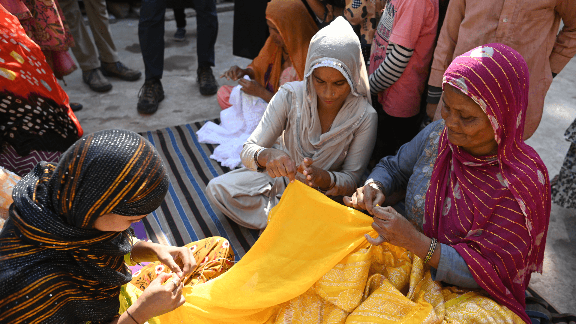 women sit together outdoors sewing and working on colorful fabric during a community gathering.