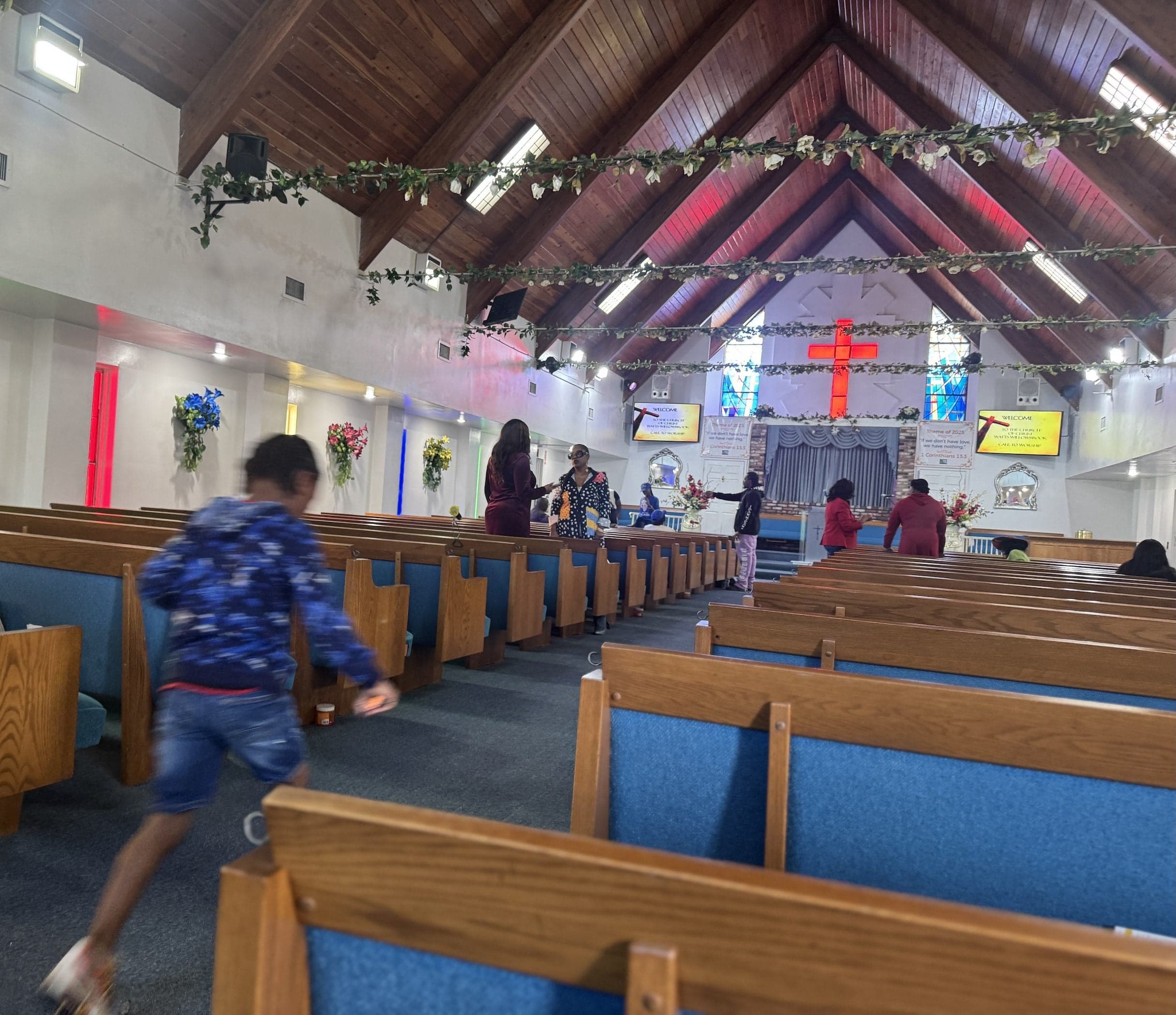 people gather inside a church sanctuary with wooden pews, stained glass windows, and a glowing cross above the altar.