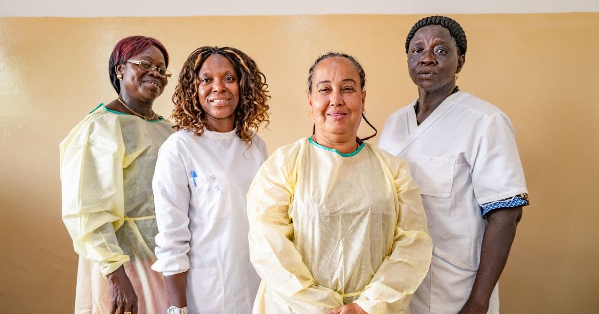 four healthcare workers standing together and smiling while wearing medical uniforms and protective gowns in a clinical setting.