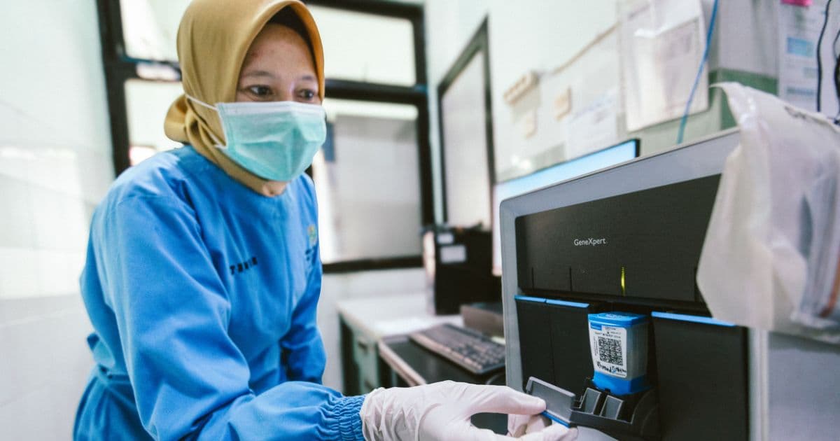 a healthcare worker wearing a mask, gloves, and protective clothing operates diagnostic testing equipment in a laboratory setting.