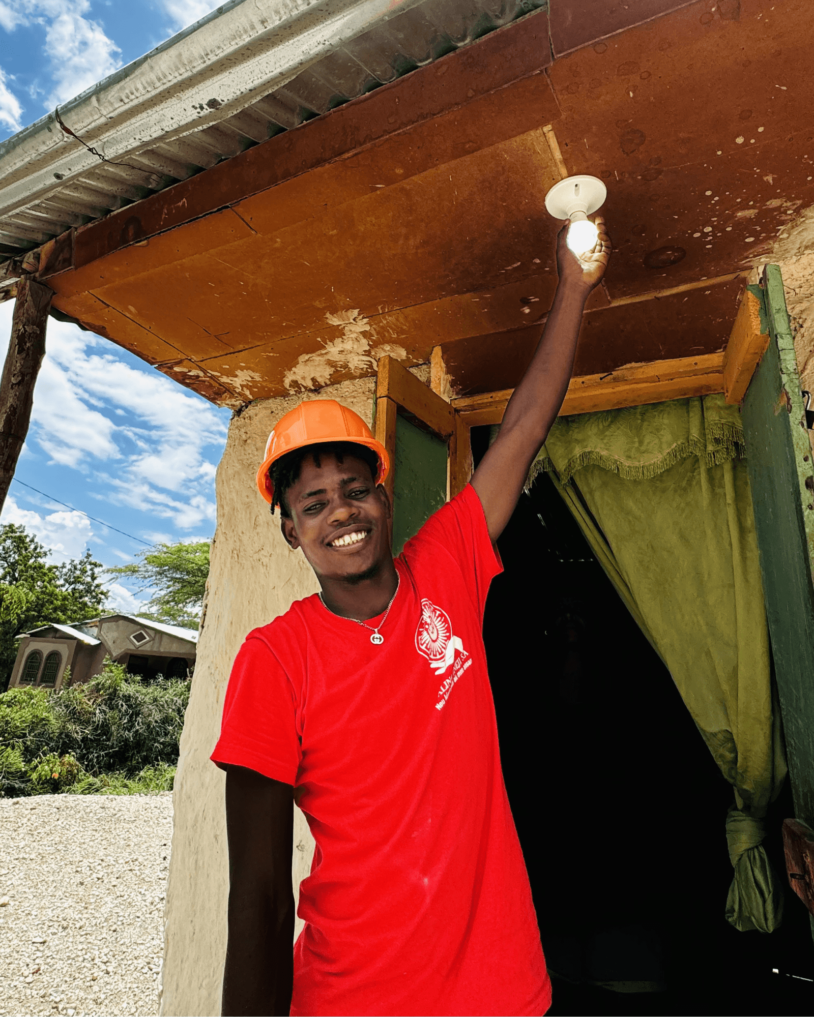 a solar energy worker wearing a hard hat smiles while turning on a newly installed light outside a home in Haiti.