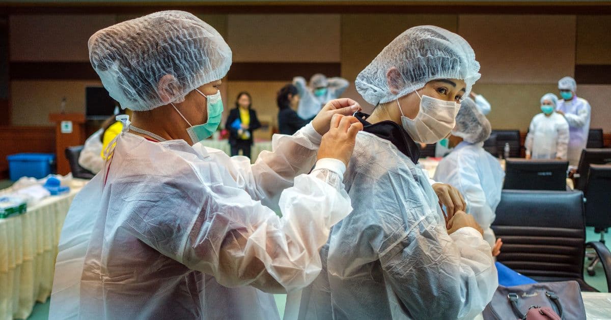 two healthcare workers wearing masks, hair covers, and protective gowns adjust personal protective equipment during a medical training session.