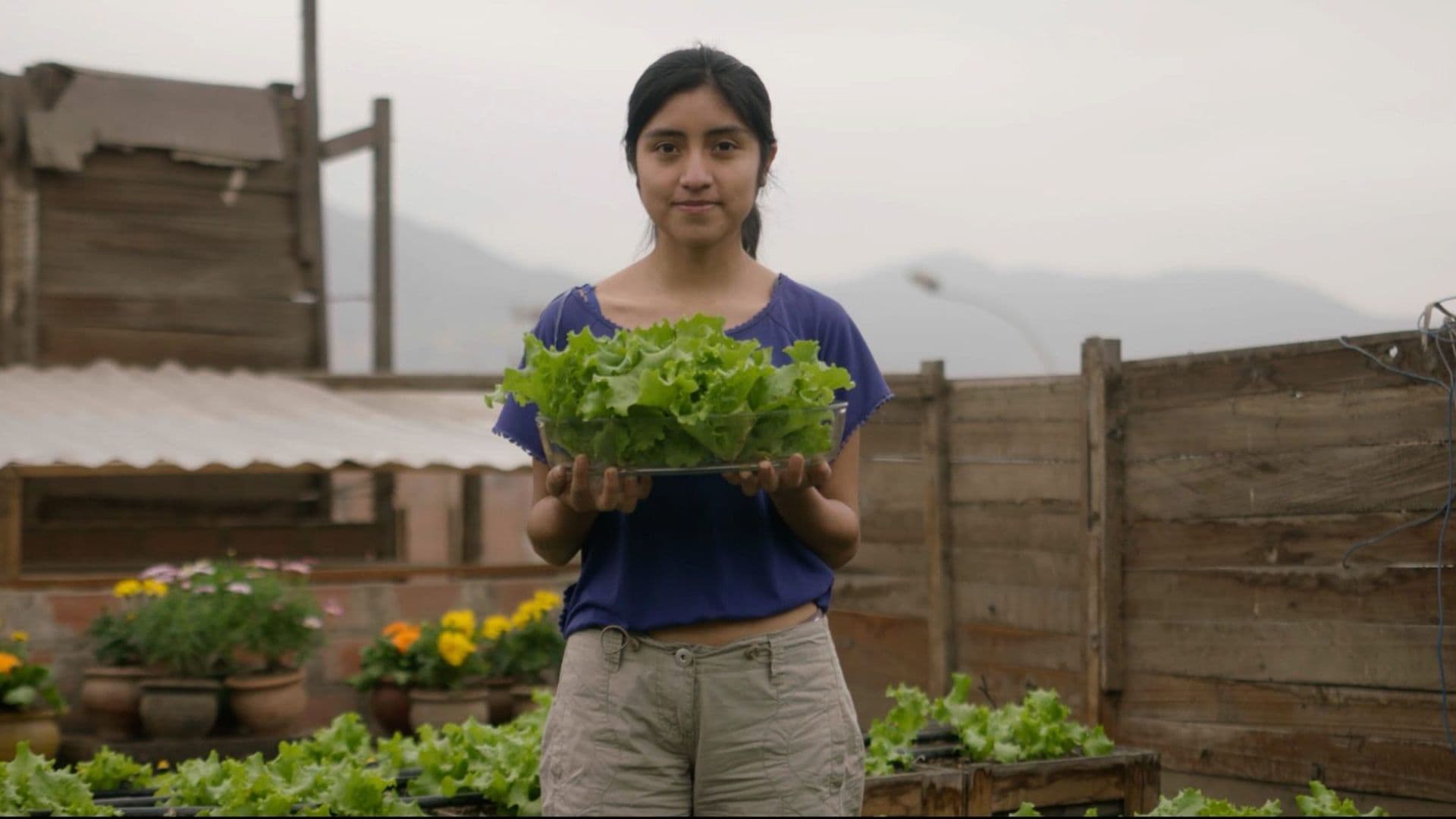 young woman standing in a community garden holding freshly harvested lettuce, with raised garden beds and flowers visible in the background.