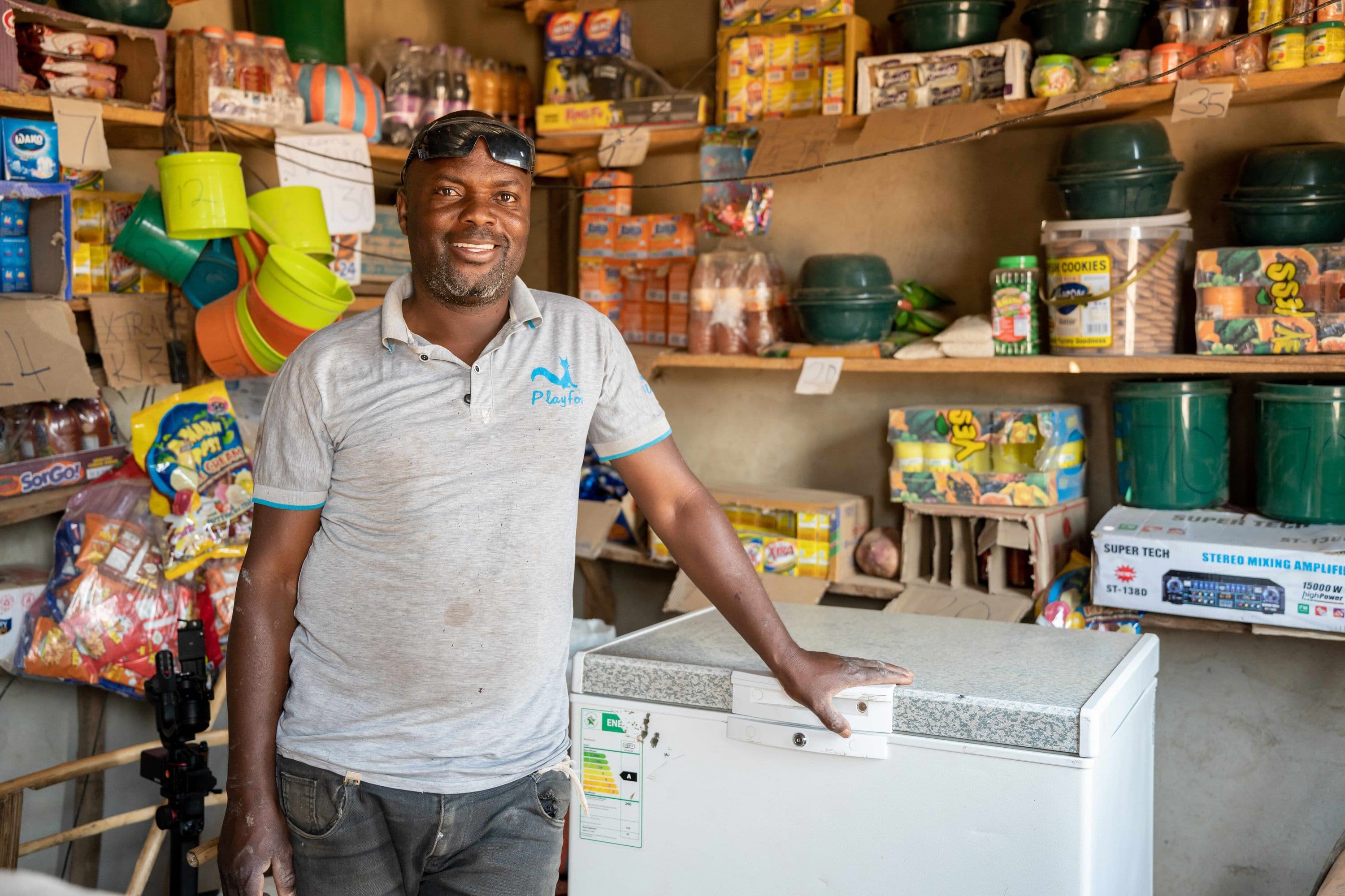 a shop owner stands inside a small store beside a chest freezer stocked with household goods and packaged food products.