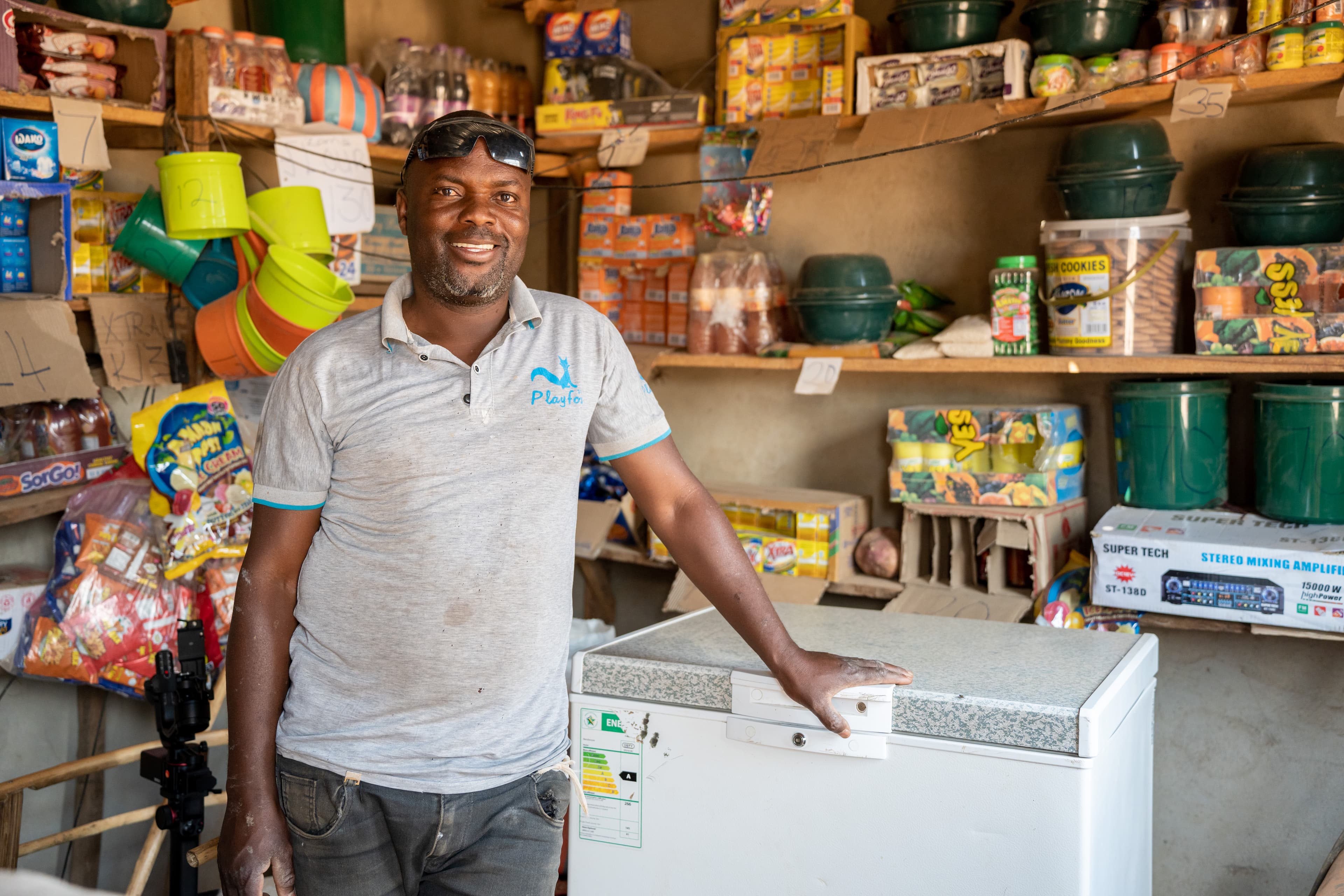 a shop owner stands inside a small store beside a chest freezer stocked with household goods and packaged food products.