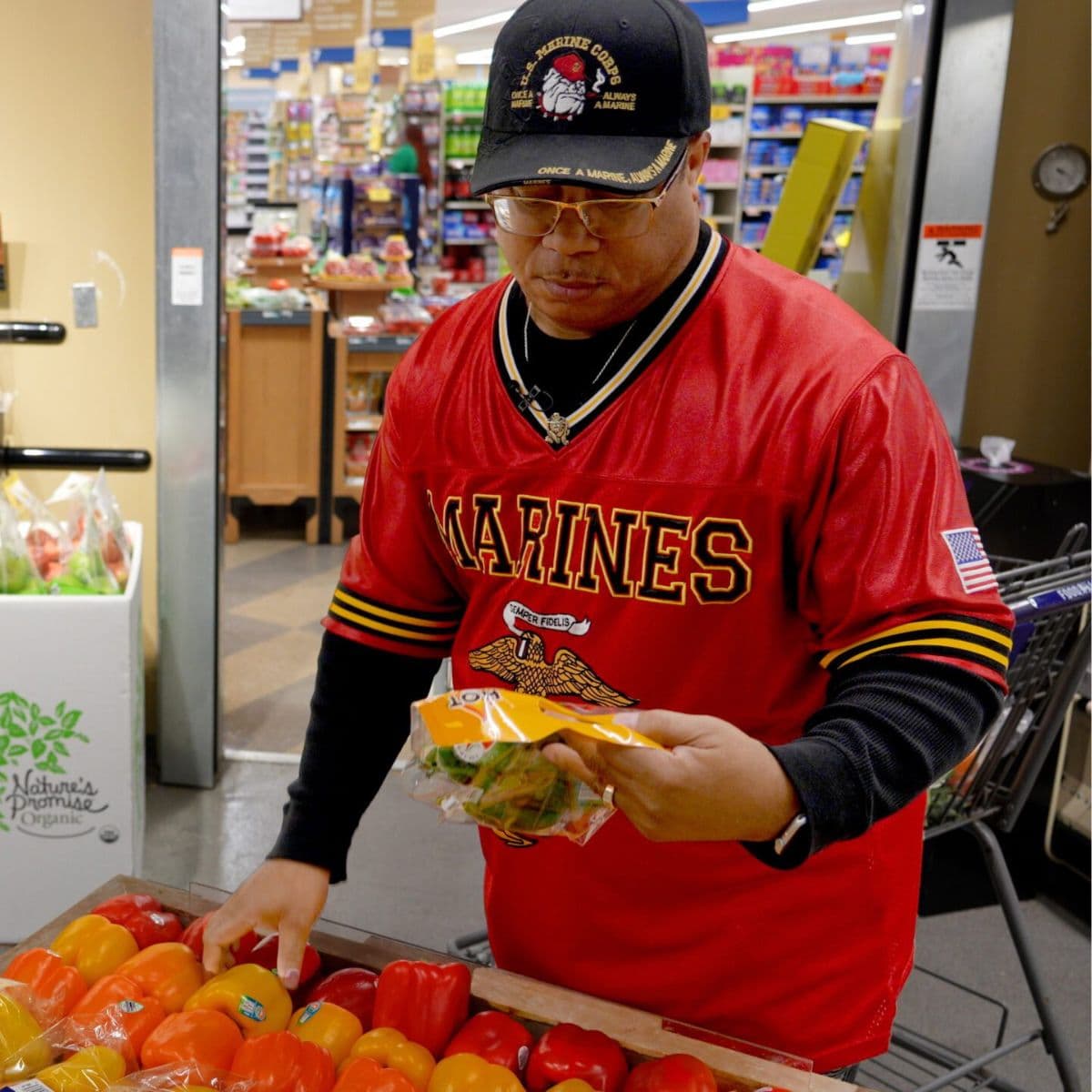 a man wearing a red Marines jersey and veteran cap selects fresh produce from a grocery display while holding a packaged salad inside a supermarket.