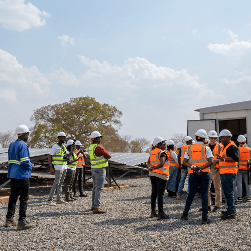 a group of workers and visitors wearing hard hats and reflective safety vests stand beside solar panels at a renewable energy site.