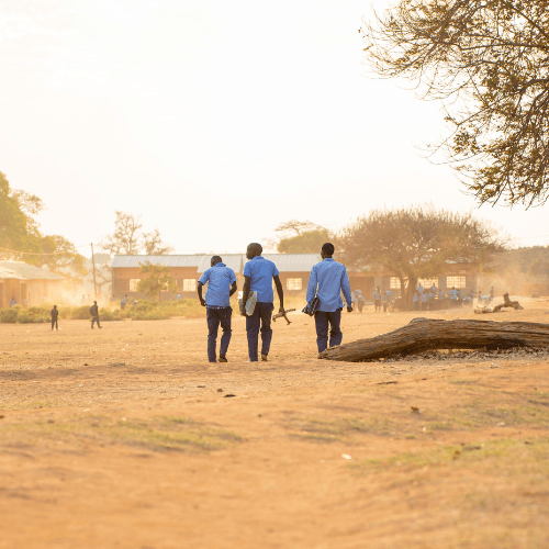 three students walk across a dusty field toward school buildings during the late afternoon.