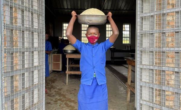a student in a blue school uniform carries a large bowl on their head while standing in a doorway.