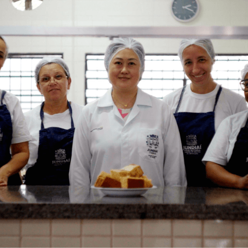 kitchen staff wearing aprons and hairnets stand together behind a counter with prepared food trays.