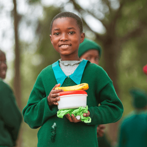 a student in a green school uniform holds a food container outdoors while smiling at the camera.