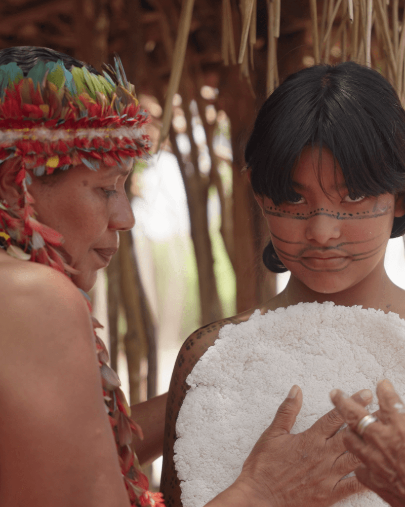 an Indigenous woman and child with traditional face paint and ceremonial attire participate in a cultural activity outdoors.