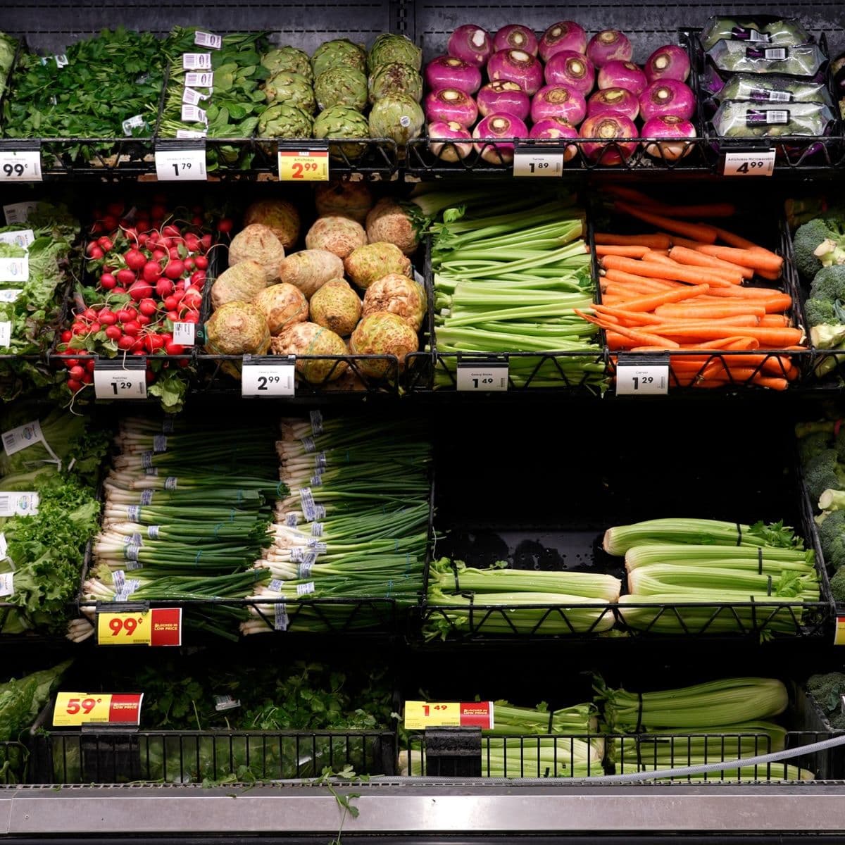 display of fresh vegetables including carrots, celery, radishes, onions, and leafy greens arranged on grocery store shelves.