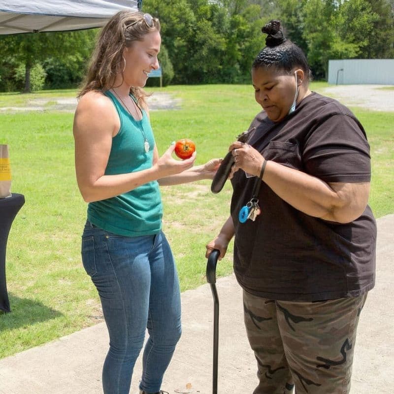 two women stand outdoors during a community food distribution event while selecting fresh vegetables, including a tomato and eggplant.