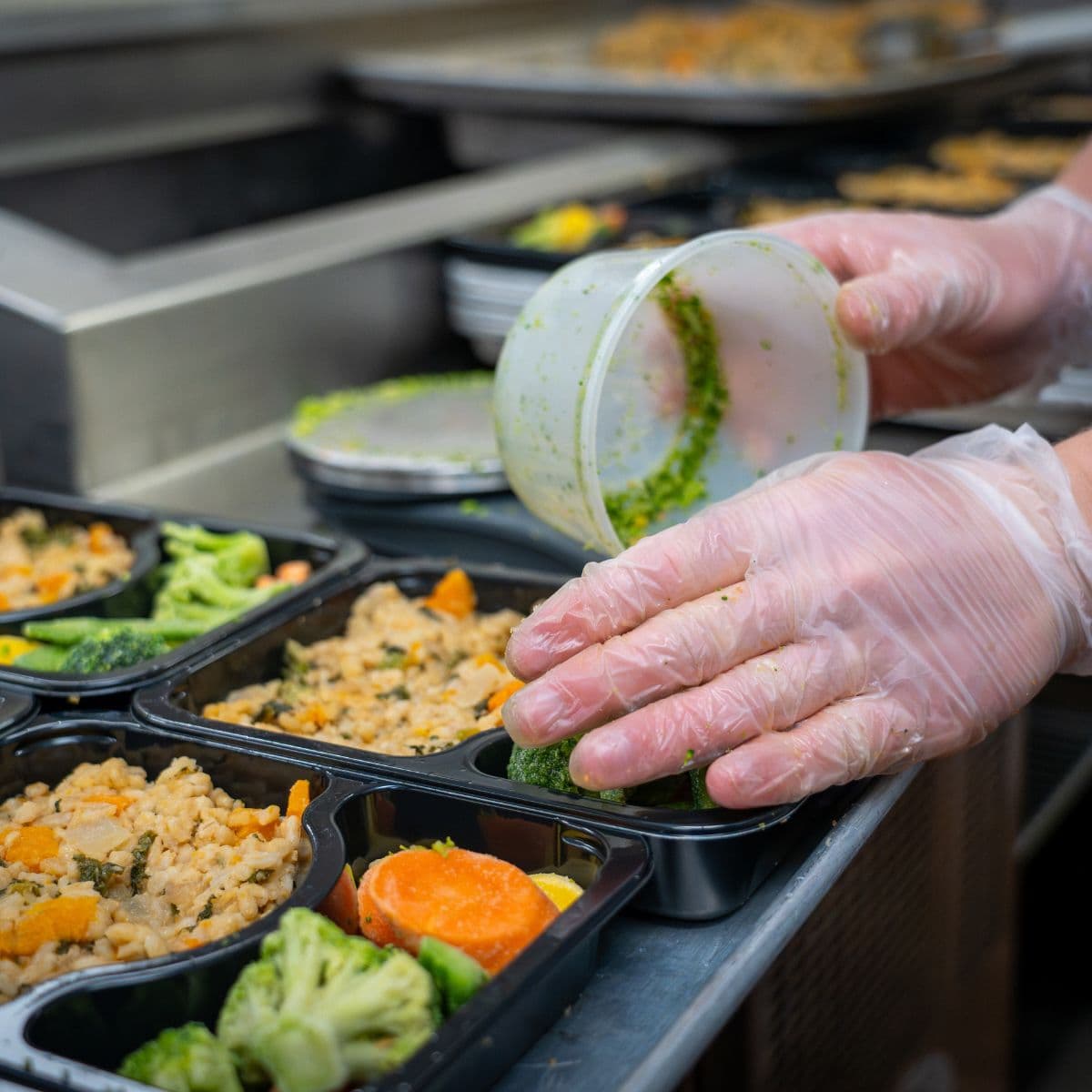 gloved hands portion vegetables and prepared food into compartment meal trays on a commercial kitchen assembly line.