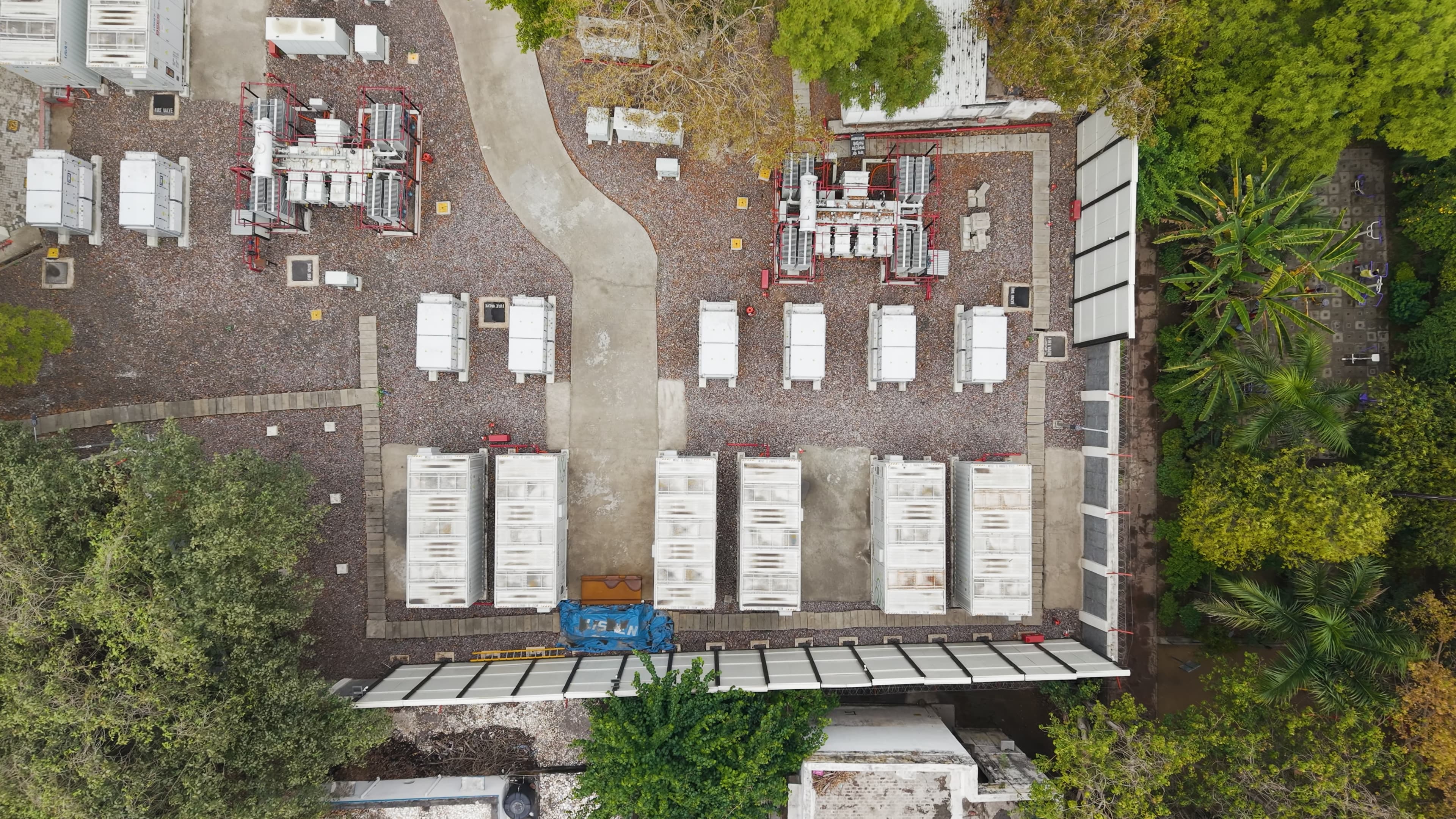 aerial view of a compact energy site with rows of battery storage units and electrical equipment arranged along a central pathway, surrounded by greenery.