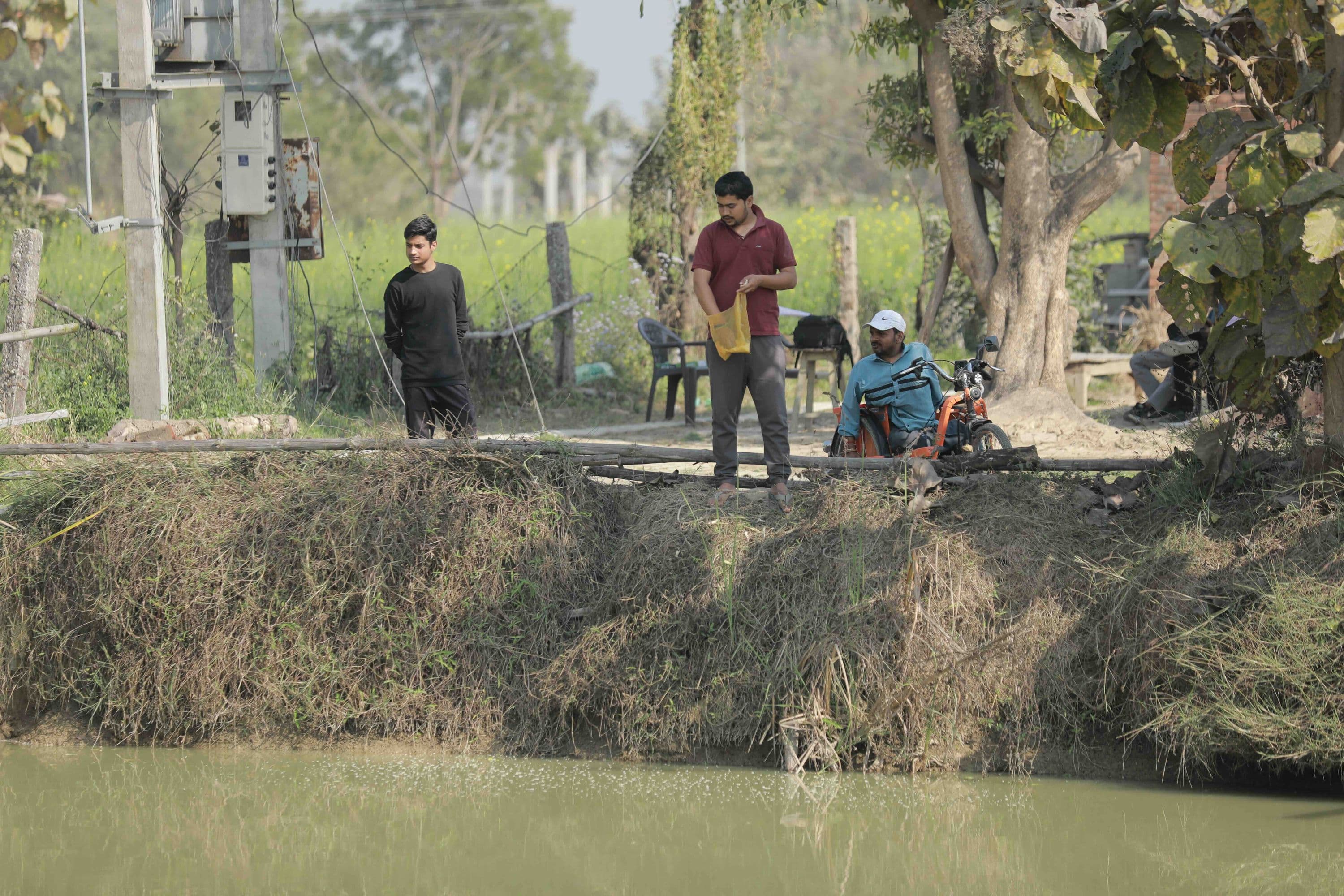 three men stand beside a pond on a rural farm, one scattering feed into the water.