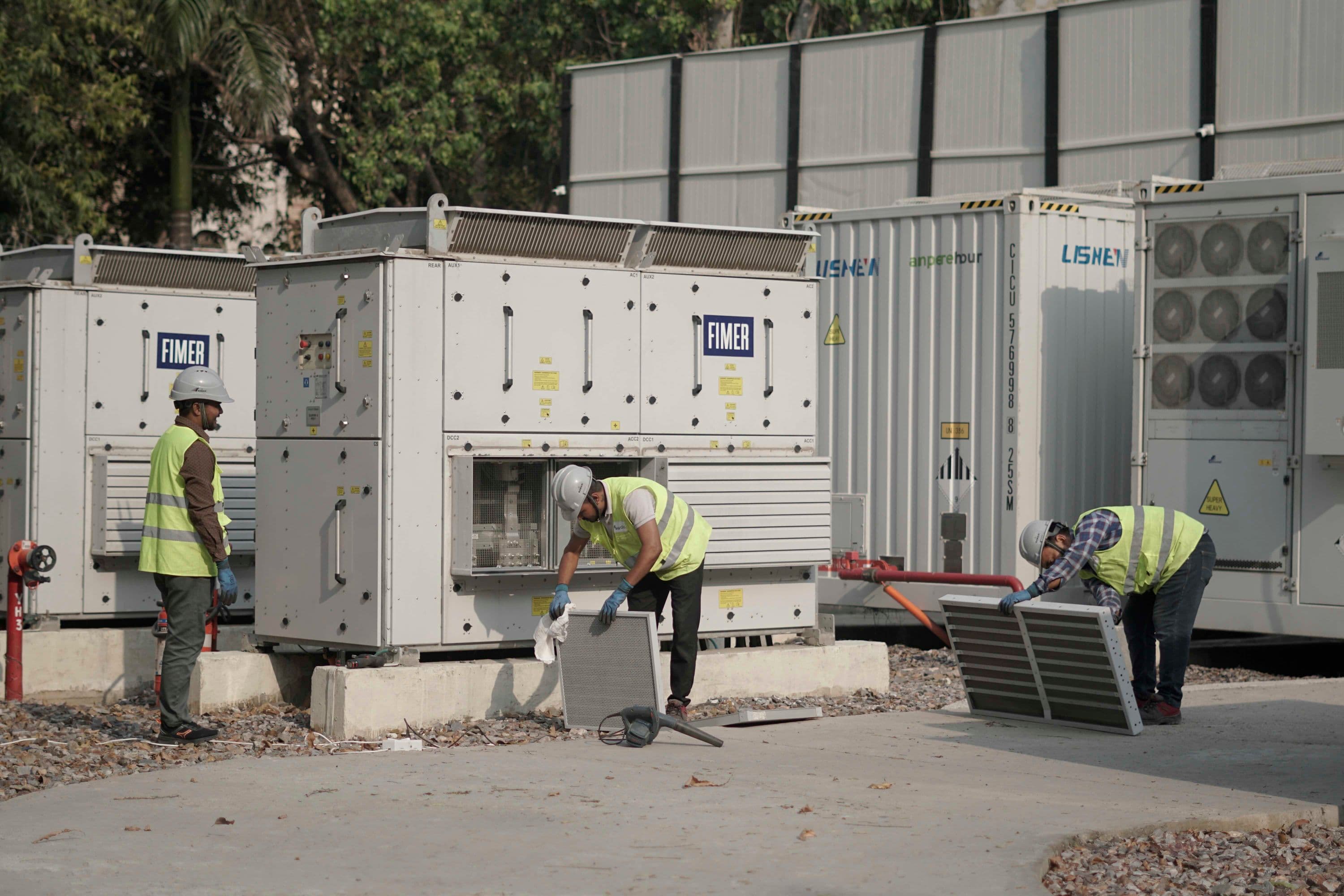 three workers in safety gear maintain battery storage units at an outdoor energy facility.